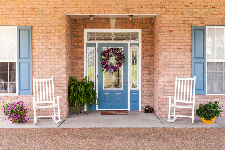 A brick front porch with a blue door, a wreath and two white rocking chairs